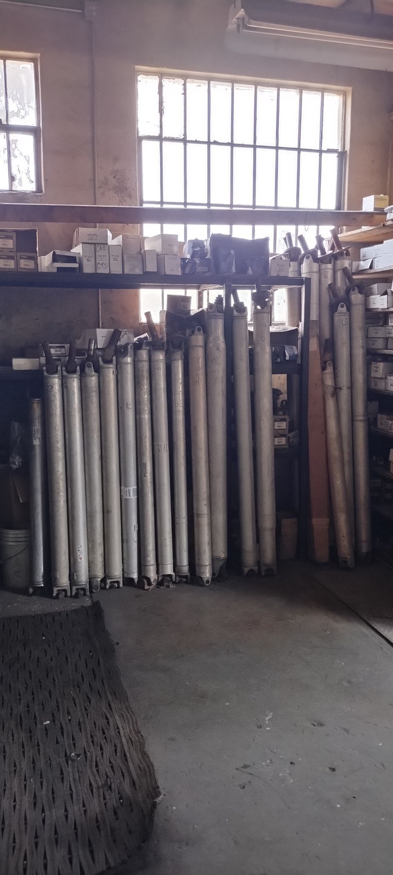A collection of metallic cylindrical driveshafts stored upright on shelves in a workshop with a window in the background.