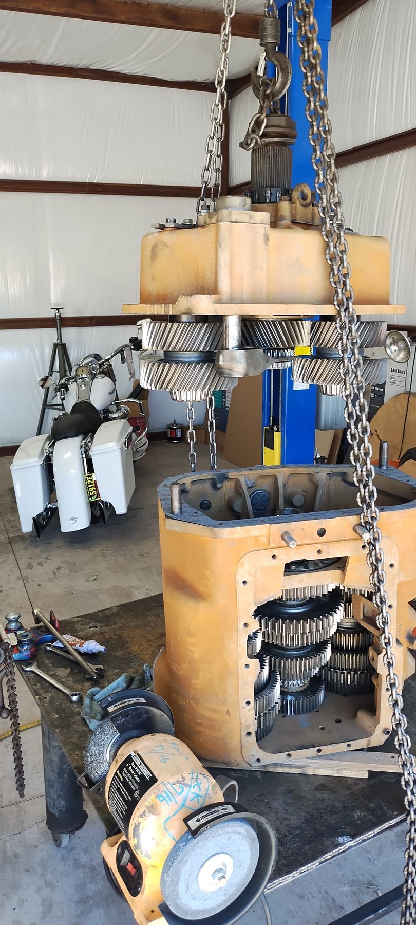 A large yellow mechanical gearbox housing being lowered by a shop crane into a work area with tools and a motorcycle.