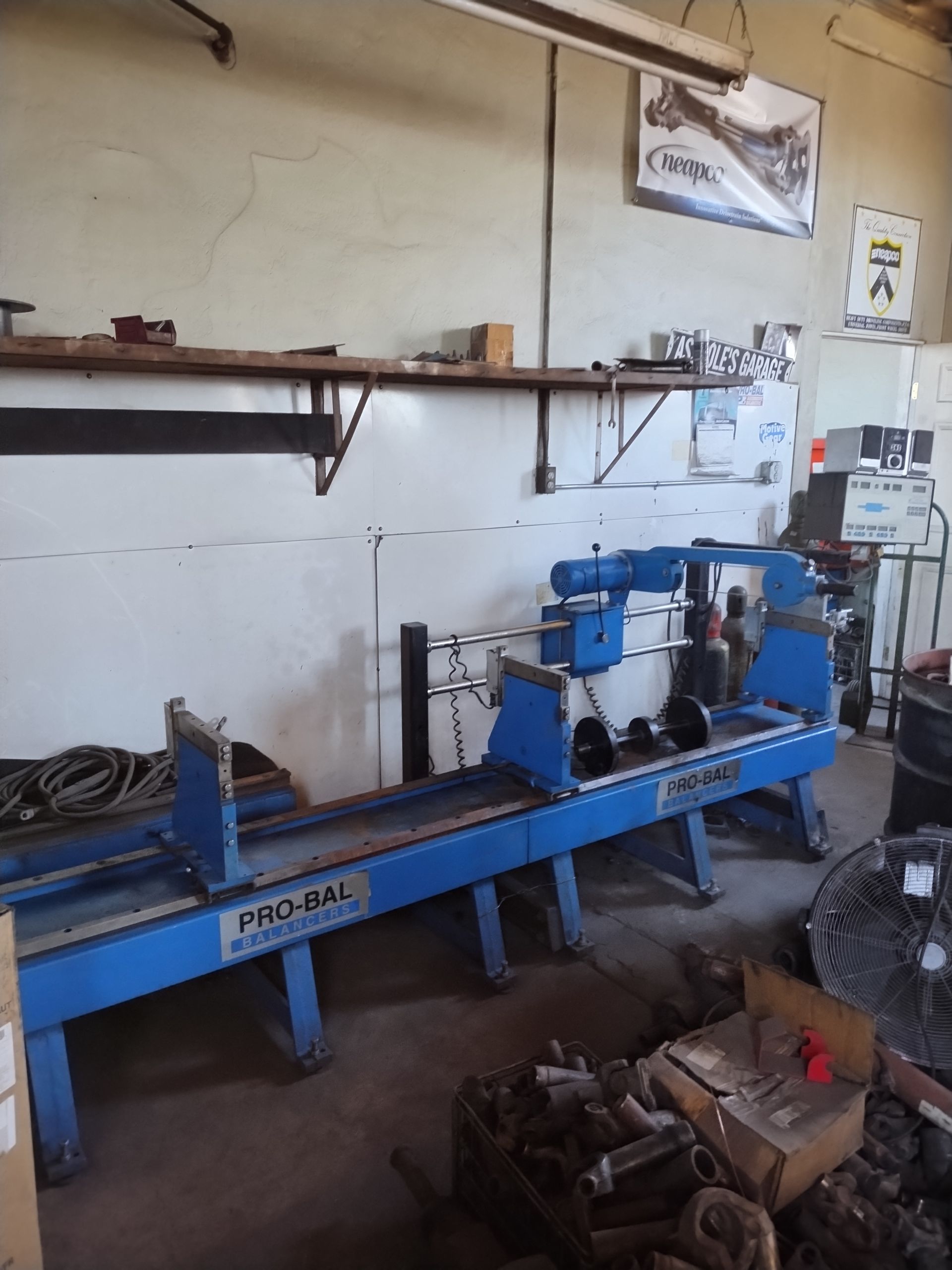 A long, blue Pro-Bal industrial machine sits on the floor of a cluttered workshop against a white wall.