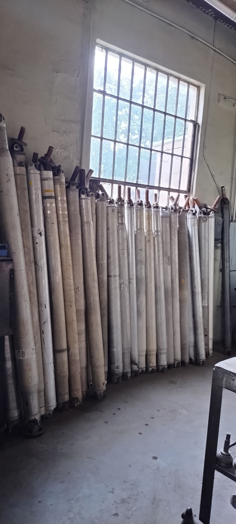 A row of metal driveshafts stands vertically against a wall in a workshop, positioned beneath a large window.