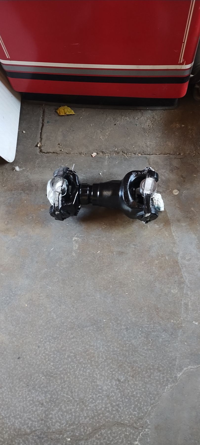 A black automotive drive shaft universal joint assembly rests on a concrete floor in front of a red structure.