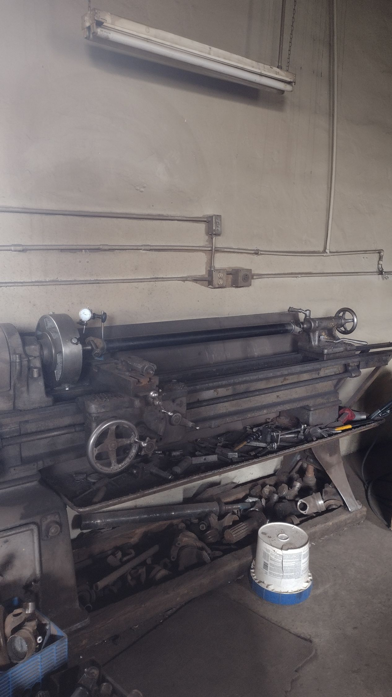 A heavy-duty metal lathe sits on a workbench in a workshop, with a white bucket on the floor nearby.