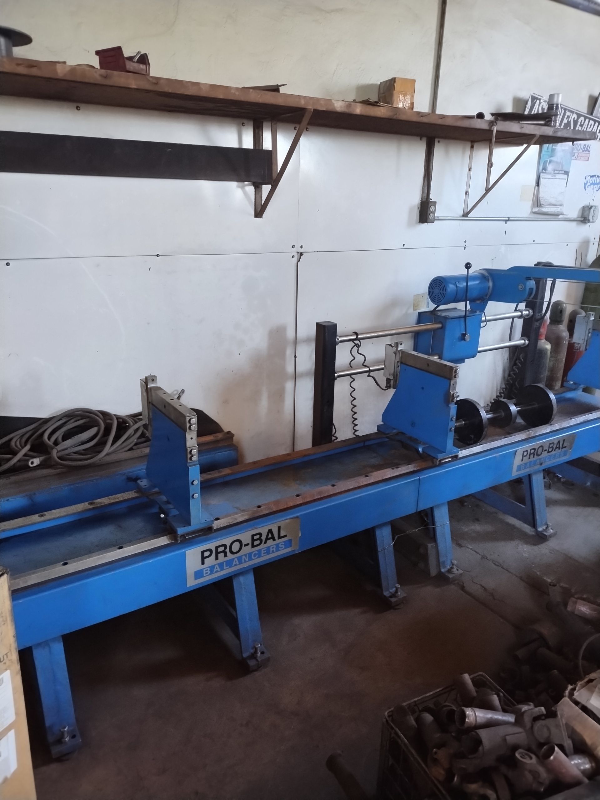 A blue Pro-Bal driveshaft balancing machine sitting on a workshop floor under a wooden shelf.