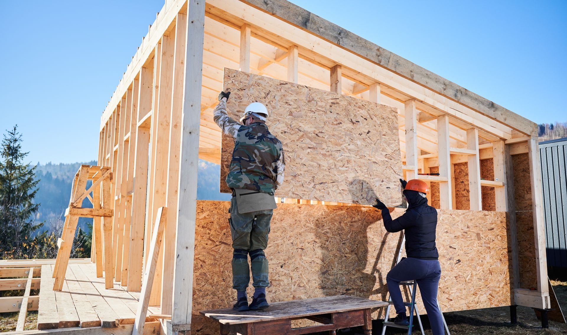Two people installing OSB sheathing on the frame of a new building in an outdoor setting.