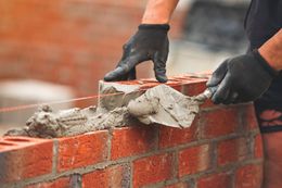 Bricklayer using a trowel to spread mortar on bricks, wearing black gloves.