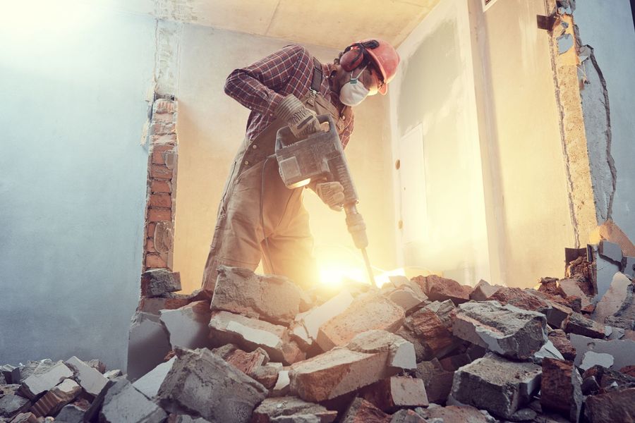 Construction worker demolishing brick wall, wearing safety gear; dusty, sunlit interior.