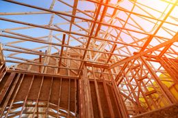 Wooden frame of a house under construction, against a bright blue sky. Sunlight streams through the beams.