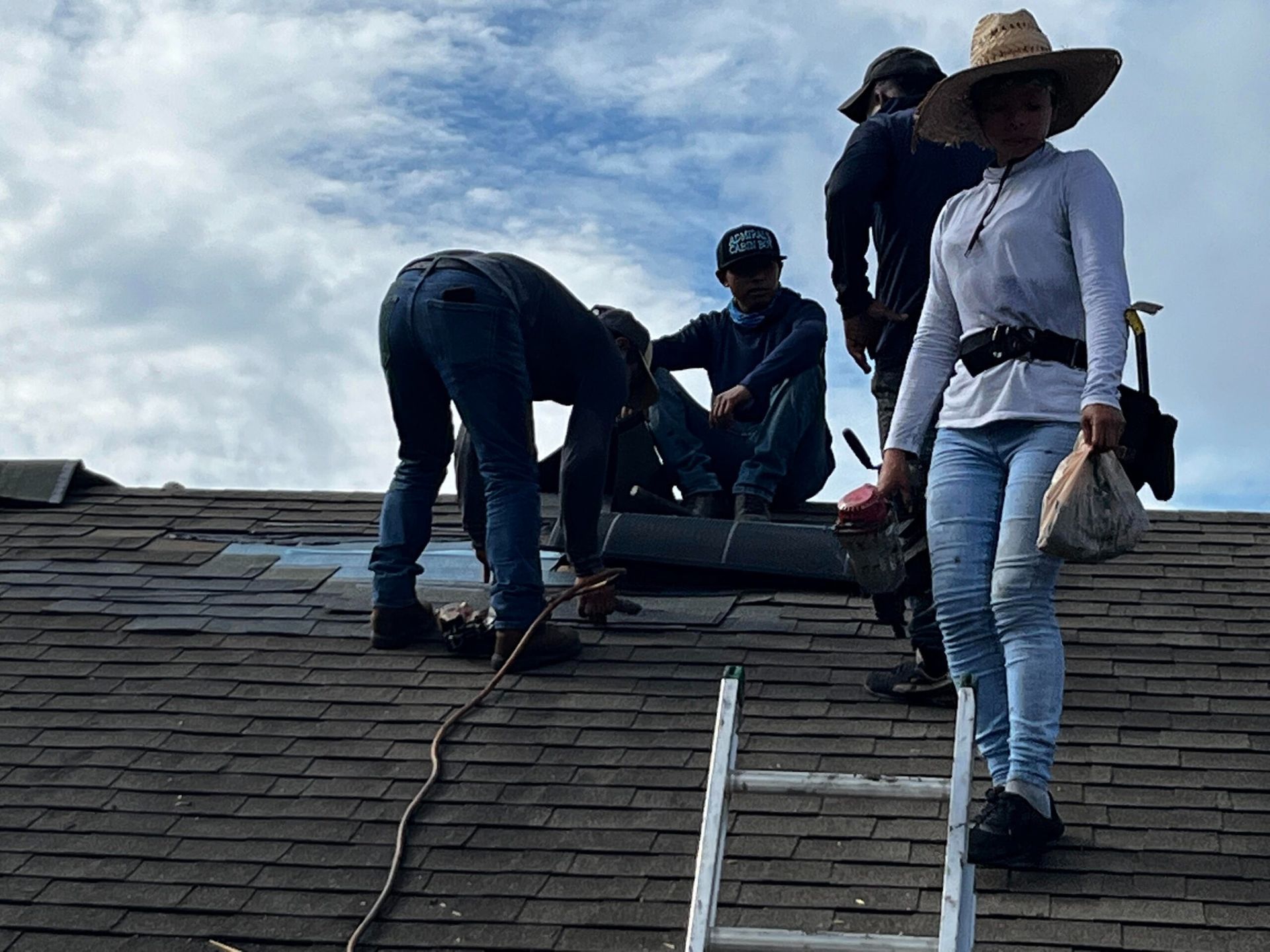 Roofers working on a dark shingled roof under a cloudy sky. One woman stands with a bag, others work.