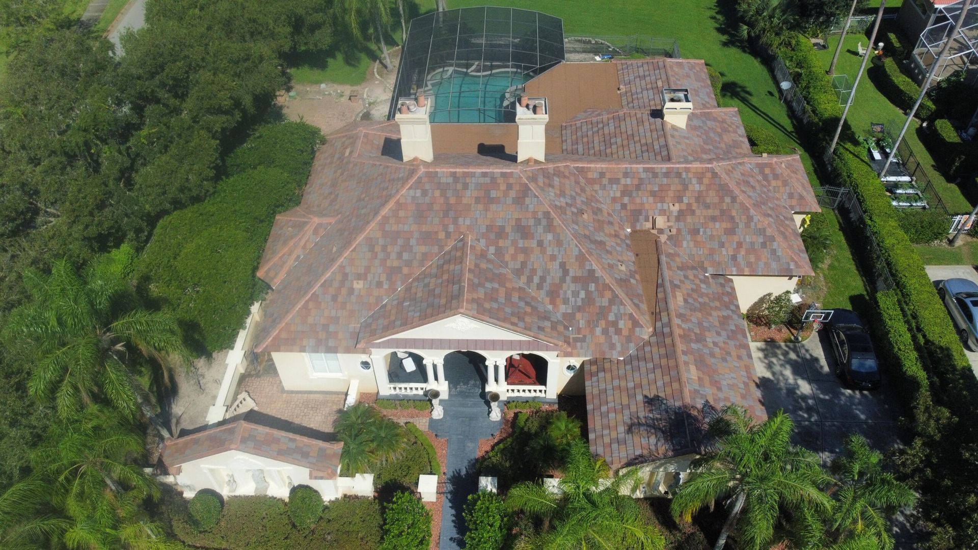 Aerial view of a large house with a brown tile roof, surrounded by lush green trees and a tennis court.