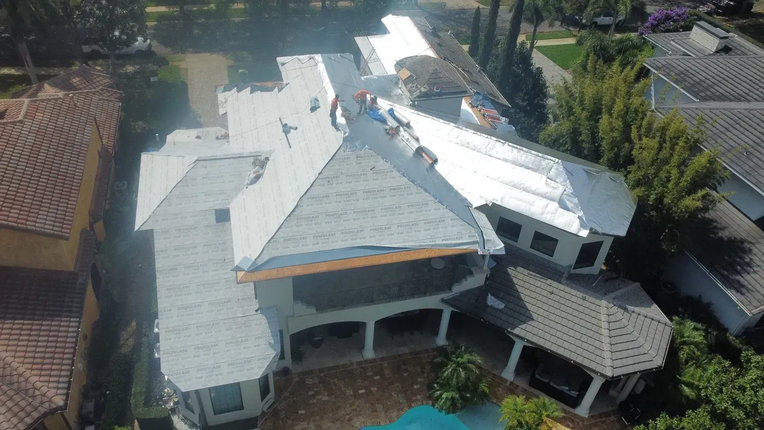 Aerial view of a large house with workers on the gray shingle roof, blue pool below.