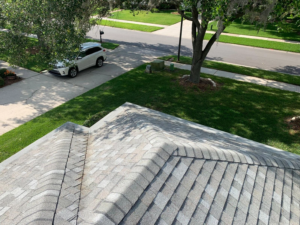 A gray shingled roof with a view of a car parked on a street with green lawns and trees.