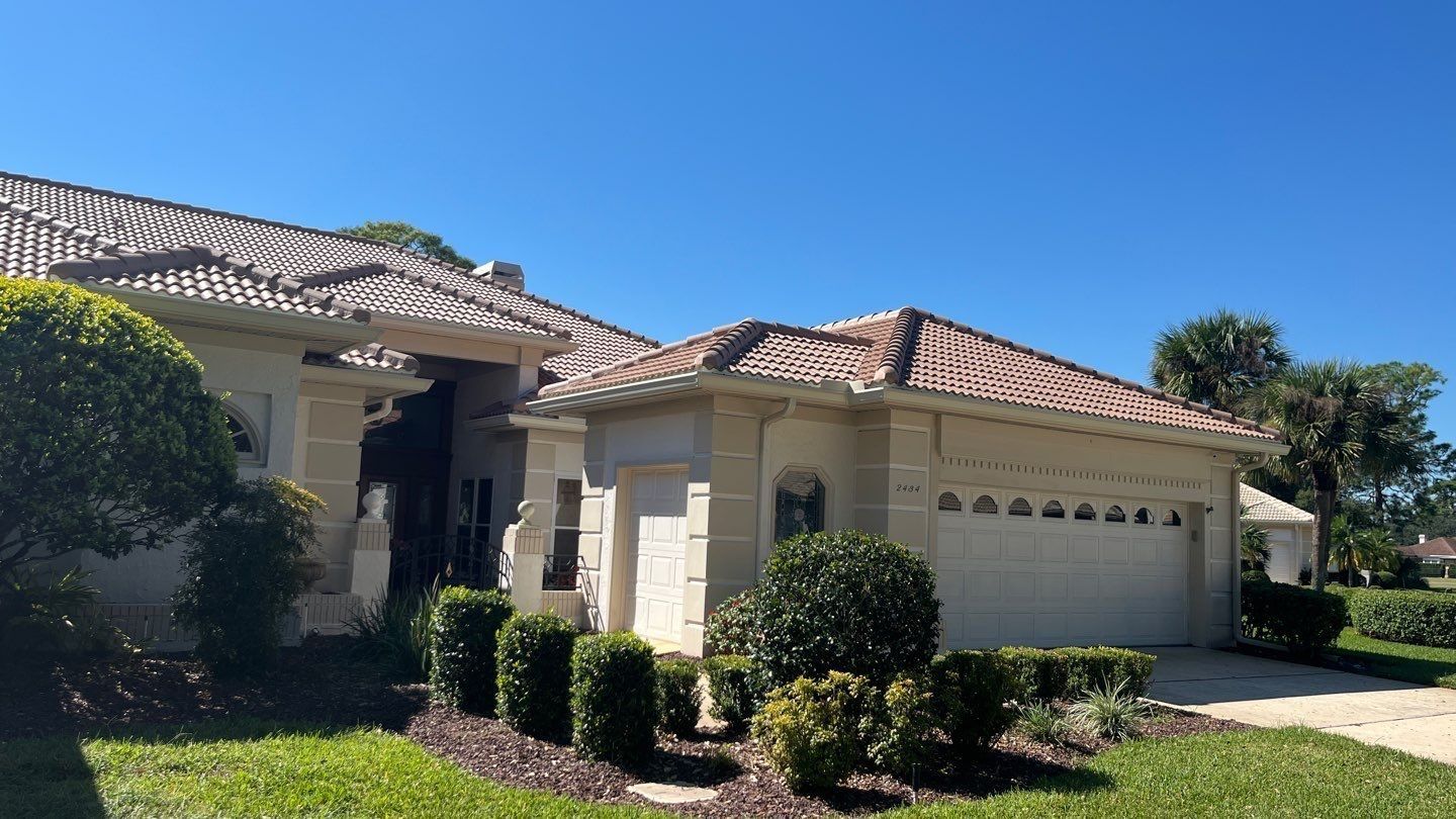 Beige house with terracotta roof and garage doors under a blue sky. Landscaping in front.