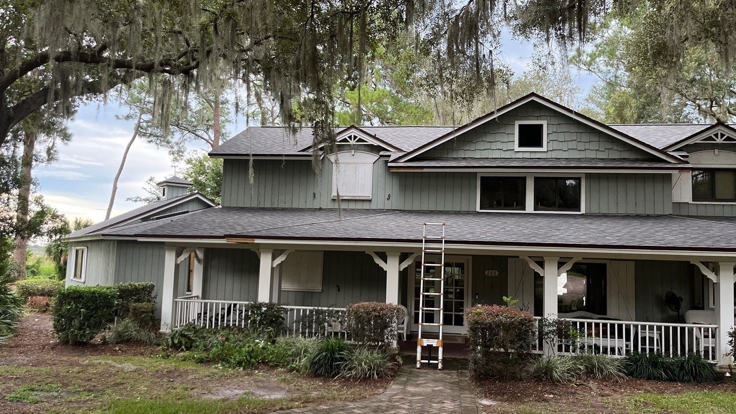 Green house with white porch and ladder, under trees.