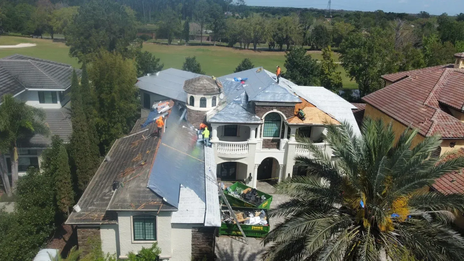 Roofers work on a large, multi-level house with a partially replaced roof.