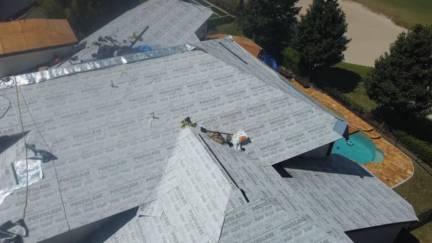 A roofing project with workers on a gray shingle roof, trees, and a pool in the background.