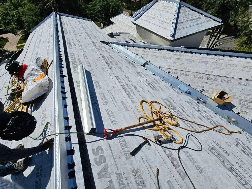 Roofer working on a gray roof, with safety harness, tools, and building materials visible.