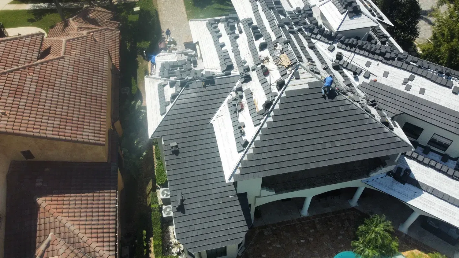 Aerial view of workers on a white roof, installing solar panels. A neighboring home with a brown roof is also visible.