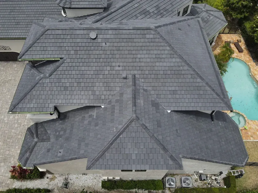 Overhead view of a gray tiled roof on a house with a pool and green trees.