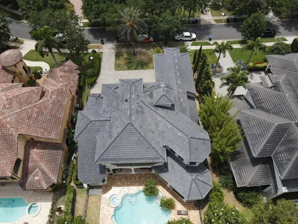 Aerial view of a large house with a pool, surrounded by trees and other houses on a sunny day.