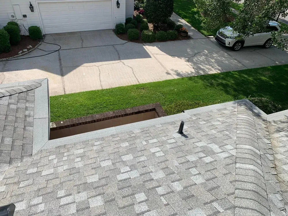 View from a roof towards a driveway, lawn, and house; gray shingles, green grass, car.