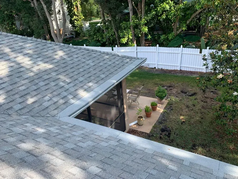 View of a roof, backyard with white fence, patio, and potted plants on a sunny day.