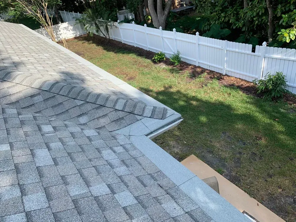 View of a gray shingle roof, lawn, white fence, and greenery under a sunny sky.