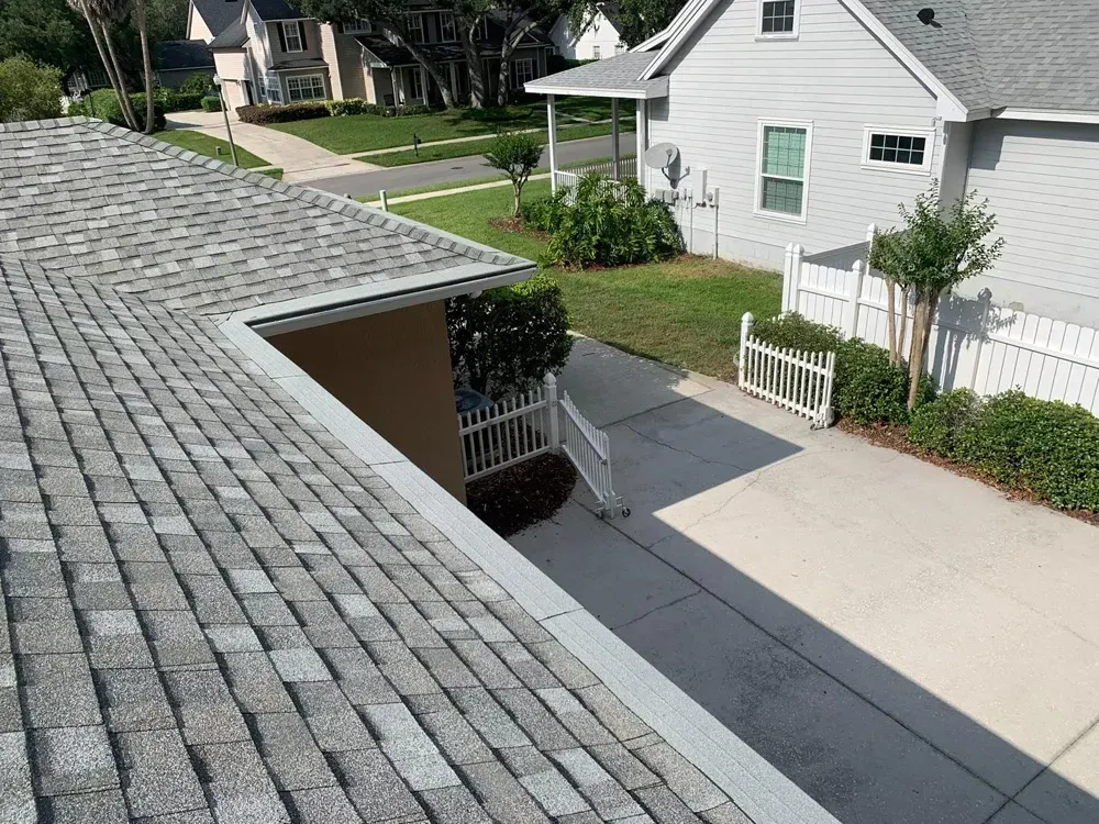 View of a gray shingled roof with a white house in the background and a driveway in front.