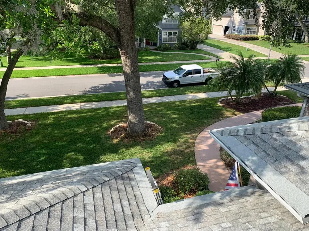 Overhead view of a house with gray roof, green lawn, street with white truck and trees in sunny neighborhood.
