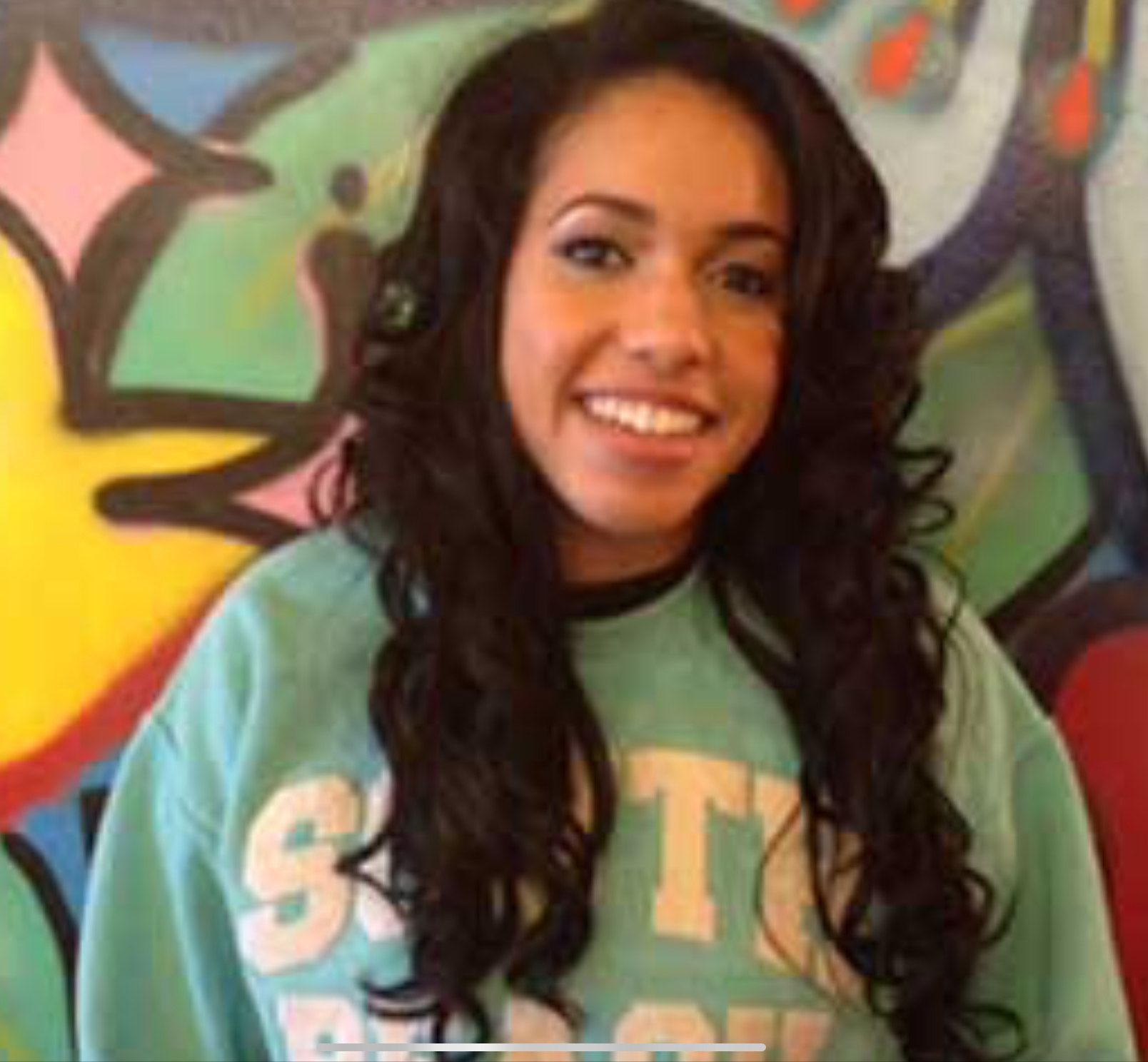 Woman with long, wavy dark hair smiles in a blue sweatshirt; graffiti art background.