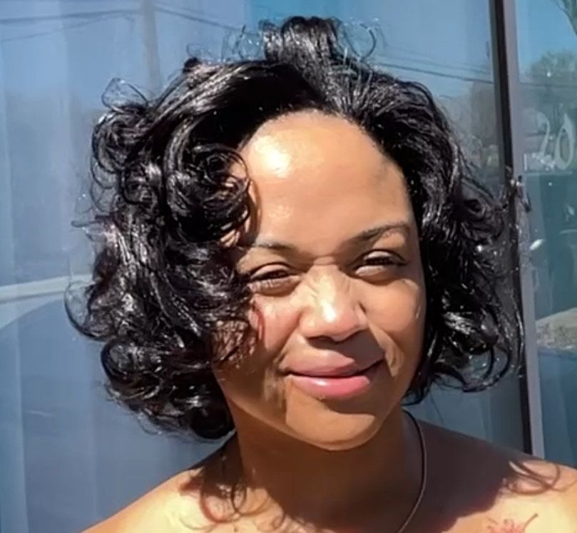 A woman with curly hair is smiling in front of a window.