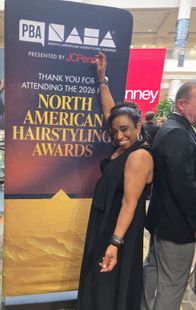Woman in a black dress posing by a North American Hairstyling Awards banner, smiling at an event.
