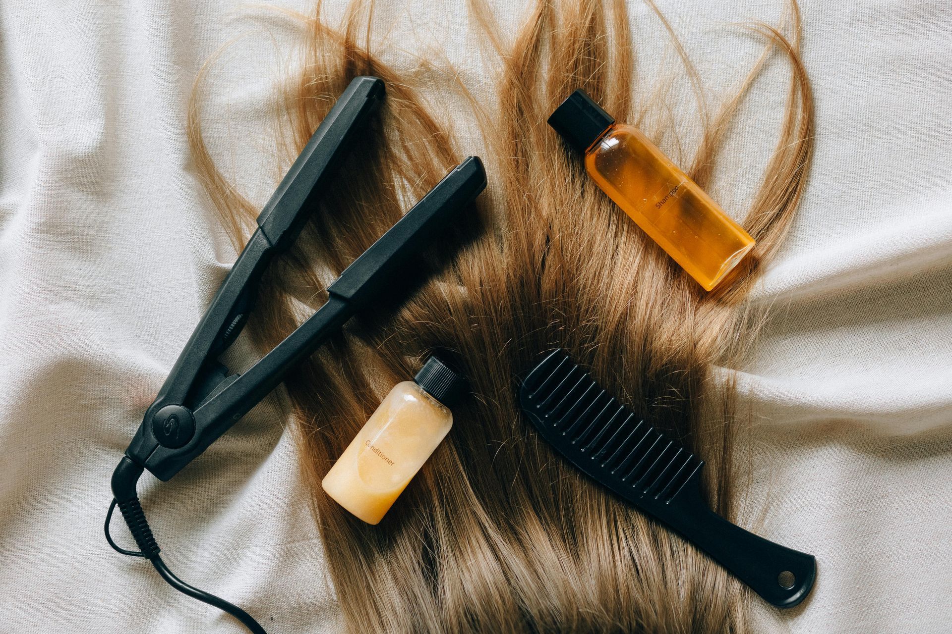 A hair straightener , comb , and bottles of hair oil are on a bed.