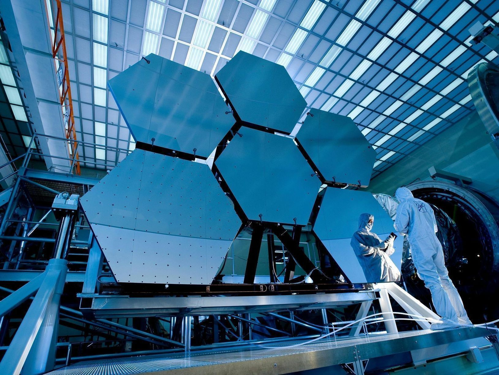 Large hexagonal mirrors assembled, being worked on by technicians in a high-tech facility. Blue and silver.