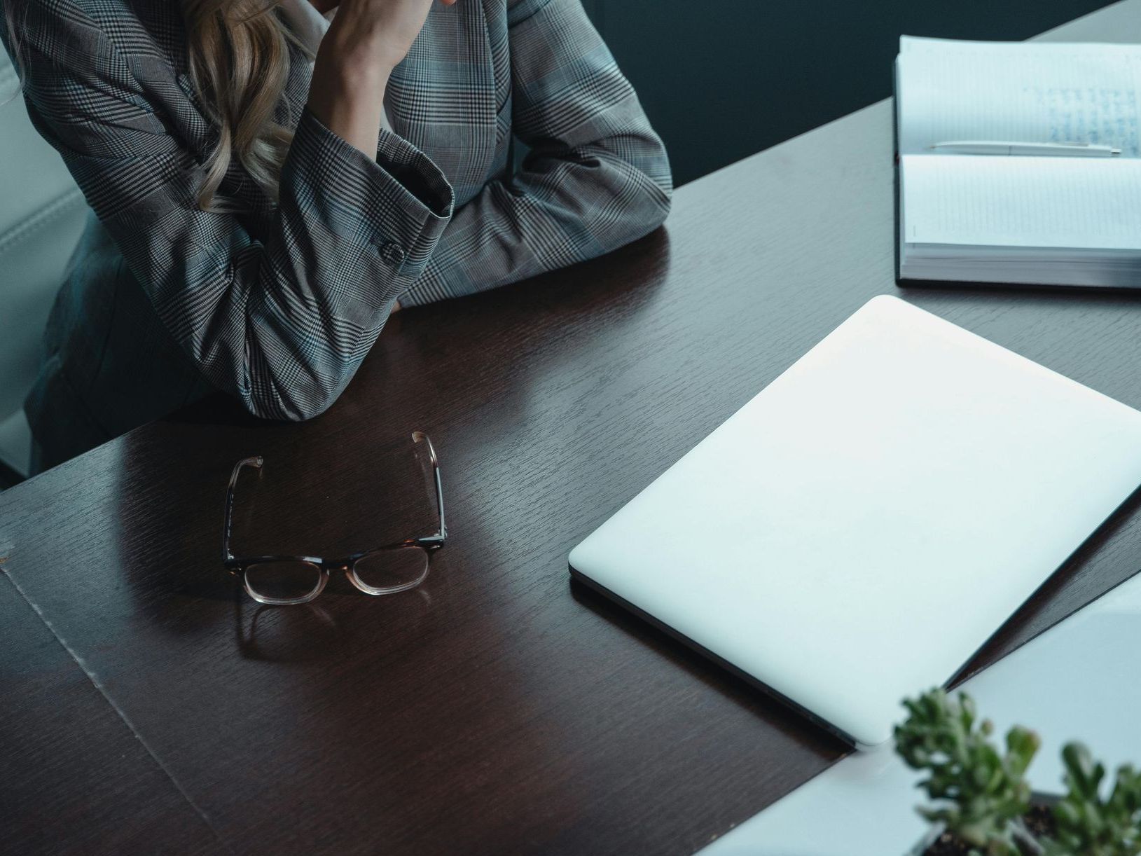 Woman at desk, chin in hand, near laptop and glasses. A notebook and small plant are also visible.