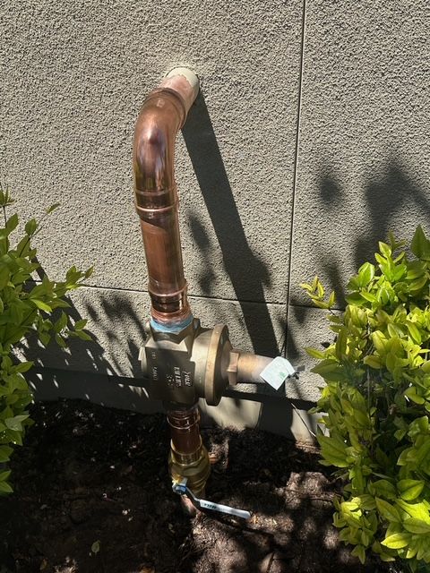 A copper pipe is attached to a wall next to a bush.