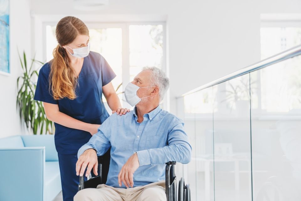 Young woman nurse explaining information to man patient in wheelchair in medical face mask while talking together in hospital.