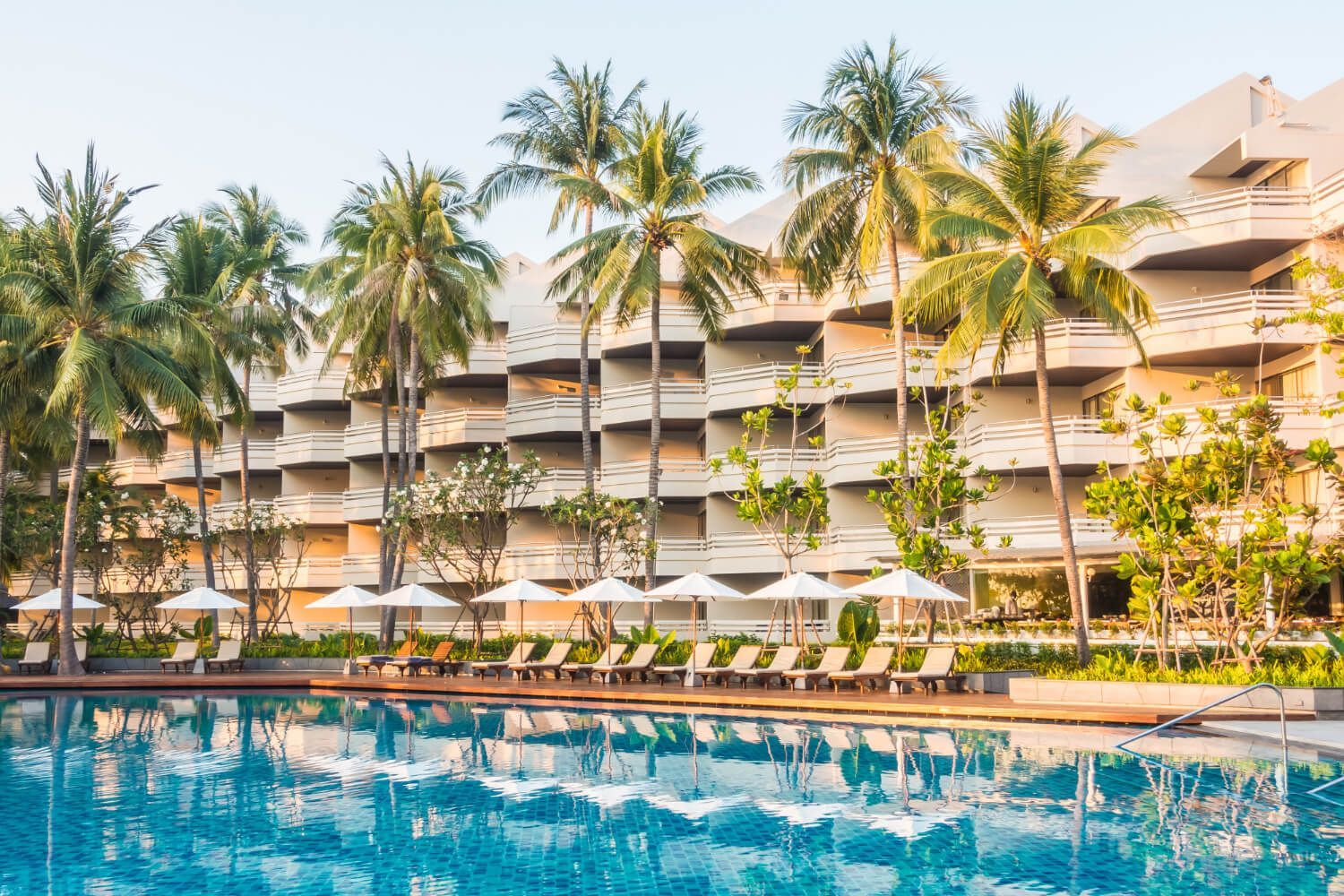 There is a large swimming pool in front of a hotel surrounded by palm trees.