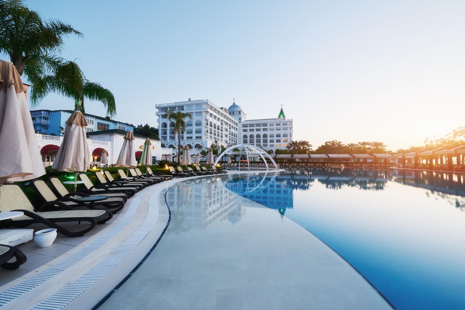 A large swimming pool surrounded by chairs and umbrellas in front of a hotel.