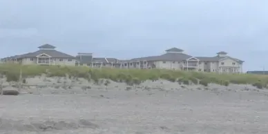 A large house is sitting on top of a sand dune on a beach.