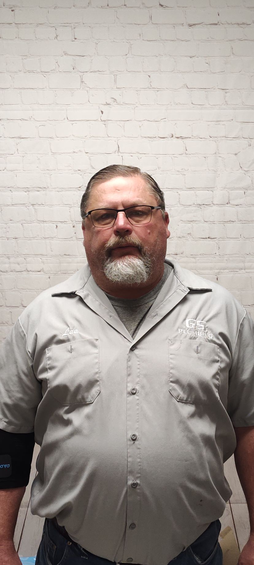 A person with glasses and a goatee stands in front of a white brick wall wearing a grey button-down shirt and a black arm.