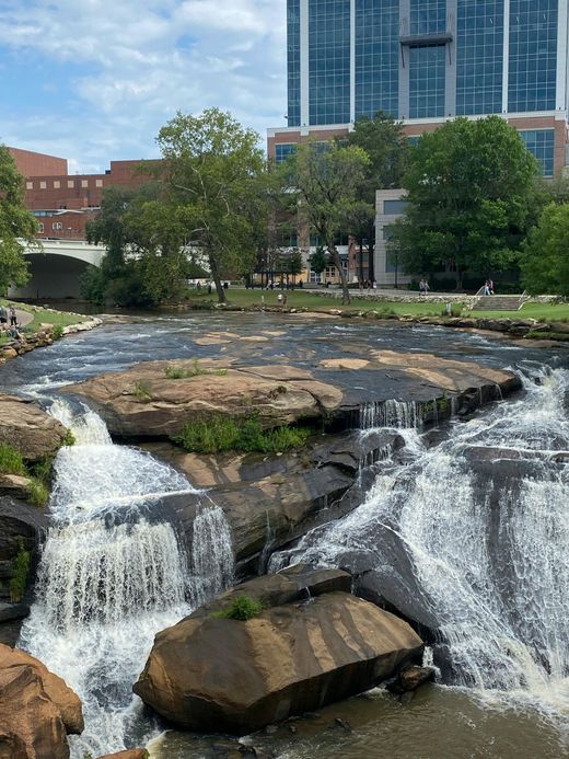 Family in kitchen: Child on step, parents cooking. Wooden cabinets, purple wall, fruits on counter, sunny.