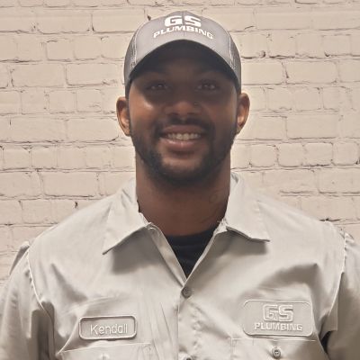 A person wearing a grey GS Plumbing work shirt and matching cap smiles against a white brick wall background.