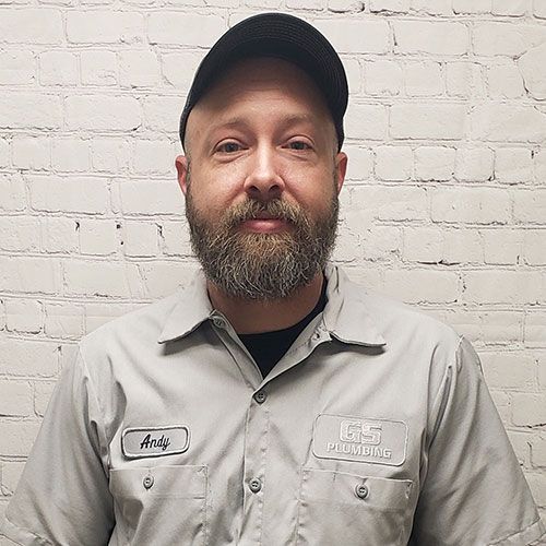 A person with a beard wearing a grey work shirt and a black cap stands in front of a white brick wall.