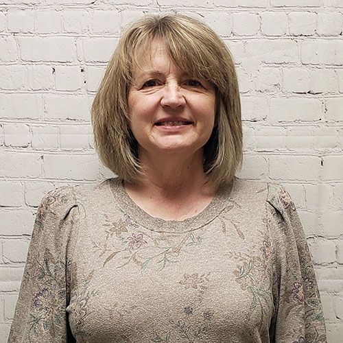 A person with shoulder-length hair wearing a long-sleeved, floral-patterned beige top, posing against a white brick wall.