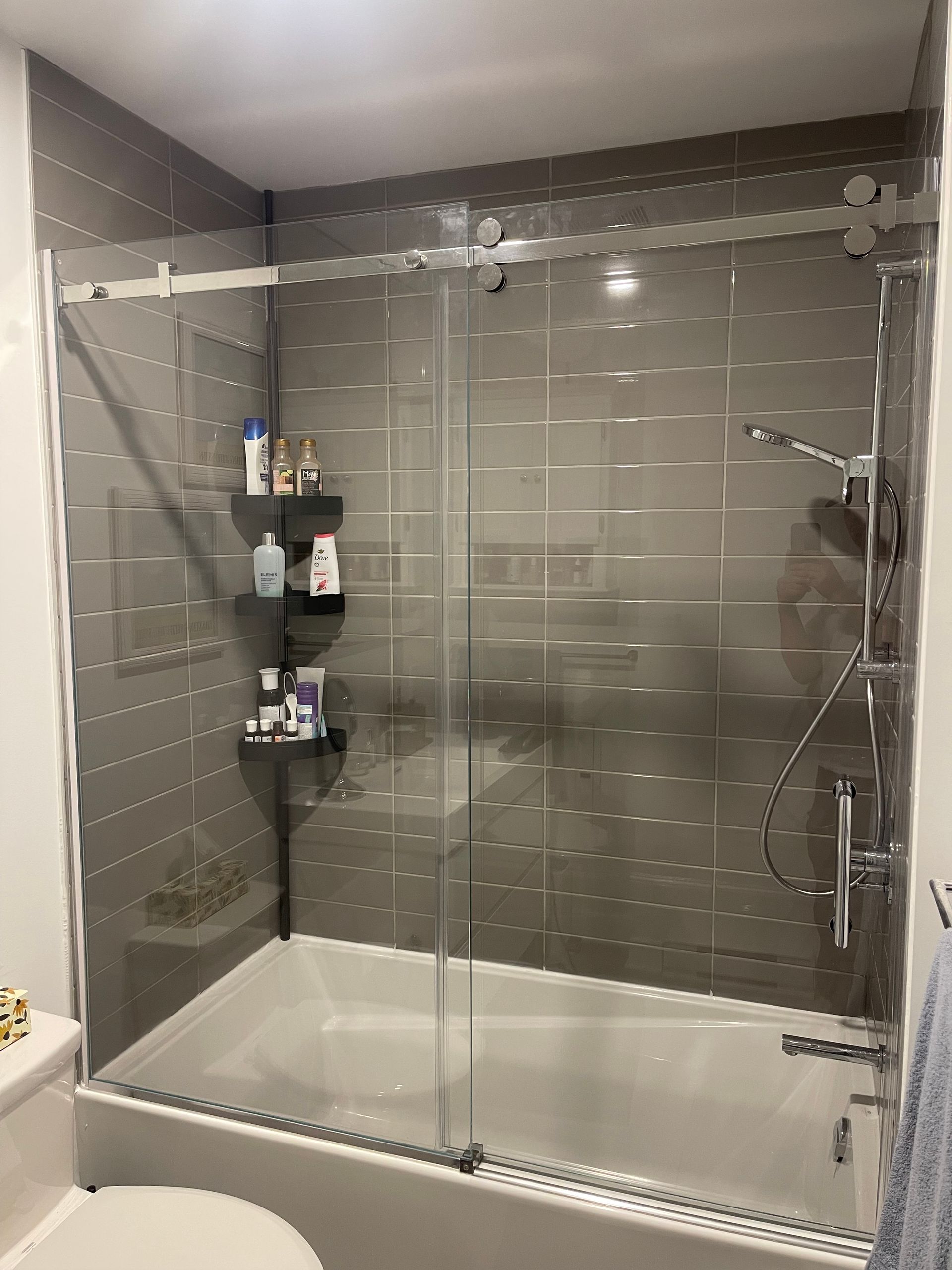 Cabine de douche carrelée grise avec portes coulissantes en verre, baignoire et articles de toilette sur des étagères.