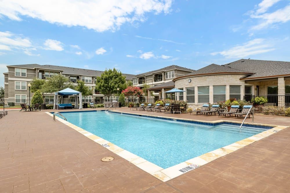 A large swimming pool surrounded by chairs and umbrellas in front of the apartment building at Marquis Rockwall in Rockwall, TX.