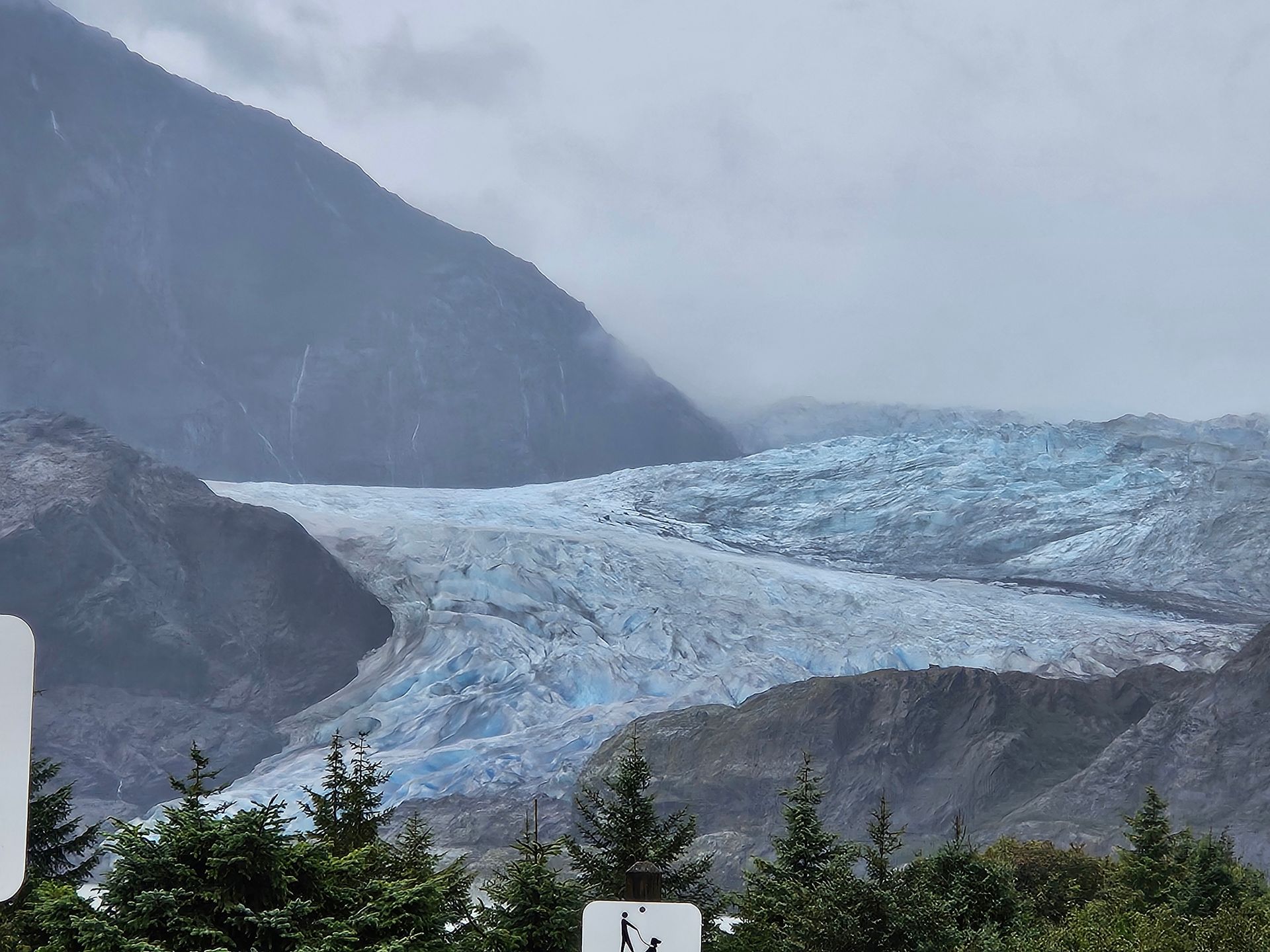 Glacier nestled in mountain range on a cloudy day, turquoise ice visible.