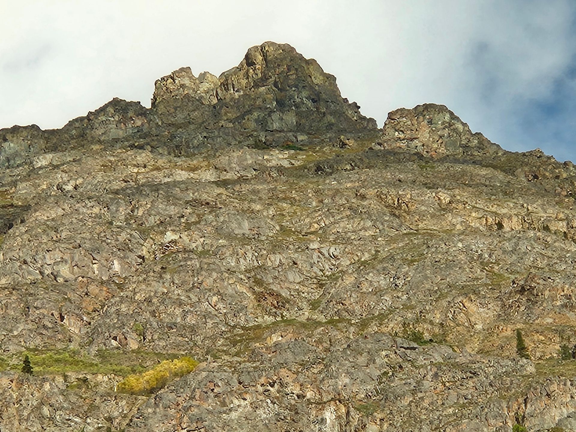 Rocky mountain peak with brown and gray rock, some green vegetation, against a blue sky.