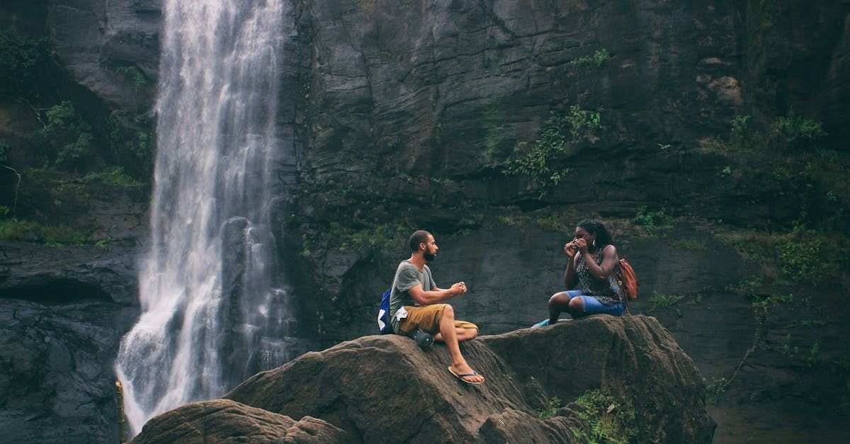 Two people sit on a rock near a waterfall, one photographing the other. Lush green foliage.