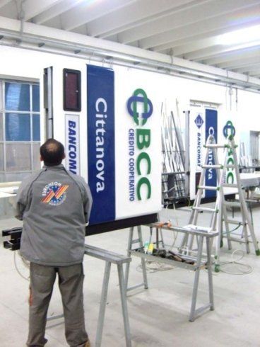 Uomo al lavoro su un grande cartello della Cittanova BCC in un'officina; colori blu e bianco.